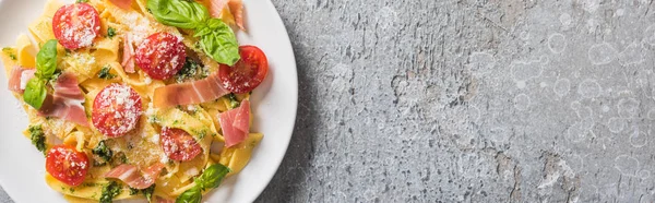 Top view of cooked Pappardelle with tomatoes, pesto sauce and prosciutto on grey surface, panoramic shot — Stock Photo