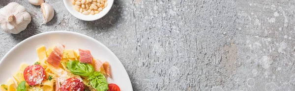 Vista dall'alto delle Pappardelle cotte con pomodori, basilico e prosciutto vicino all'aglio, pinoli su superficie grigia, colpo panoramico — Foto stock