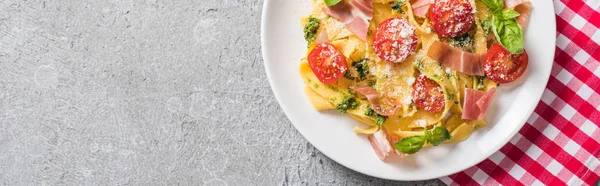 Top view of cooked Pappardelle with tomatoes, basil and prosciutto on plaid napkin on grey surface, panoramic shot — Stock Photo