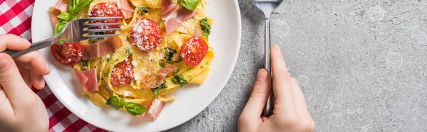 Visão parcial da mulher comer Pappardelle com tomates, manjericão e prosciutto em guardanapo xadrez na superfície cinza, tiro panorâmico — Fotografia de Stock