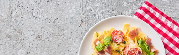 Top view of cooked Pappardelle with tomatoes, basil and prosciutto on plaid napkin on grey surface, panoramic shot — Stock Photo