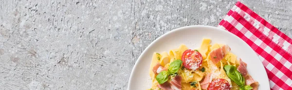 Vue du dessus de la Pappardelle cuite aux tomates, basilic et prosciutto sur serviette à carreaux sur surface grise, vue panoramique — Photo de stock