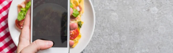 Partial view of woman taking picture of Pappardelle with tomatoes, basil and prosciutto on smartphone on grey surface, panoramic shot — Stock Photo