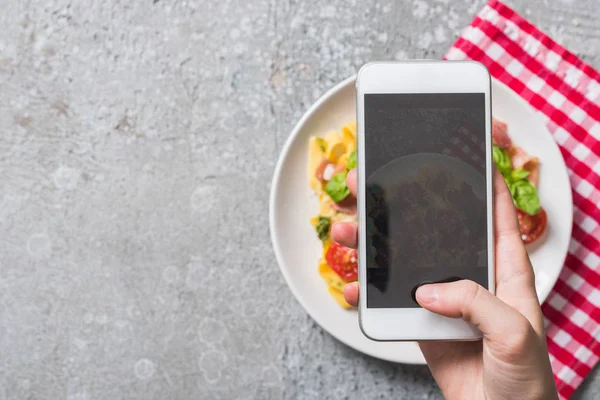 Partial view of woman taking picture of Pappardelle with tomatoes, basil and prosciutto on smartphone on grey surface — Stock Photo