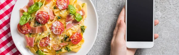 Partial view of woman holding smartphone near Pappardelle with tomatoes, basil and prosciutto on plaid napkin on grey surface, panoramic shot — Stock Photo