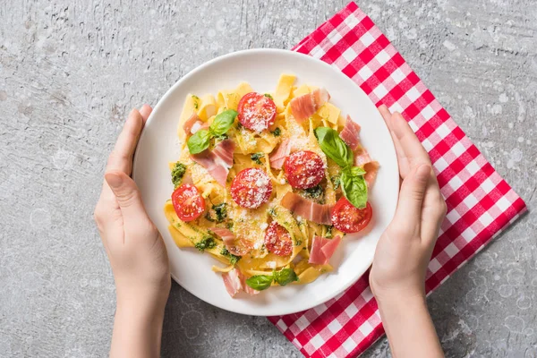 Partial view of woman holding plate with Pappardelle with tomatoes, basil and prosciutto on plaid napkin on grey surface — Stock Photo