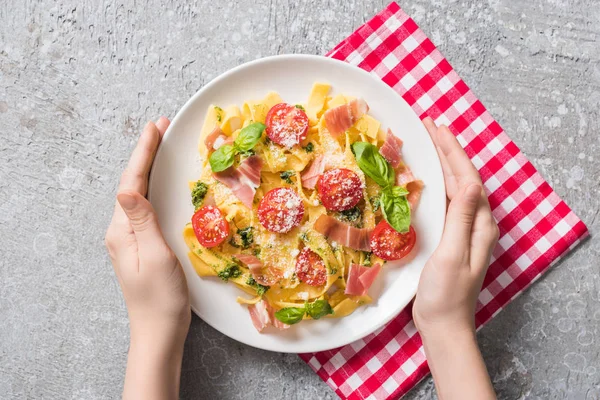 Visão parcial da mulher segurando placa com Pappardelle com tomates, manjericão e prosciutto em guardanapo xadrez na superfície cinza — Fotografia de Stock