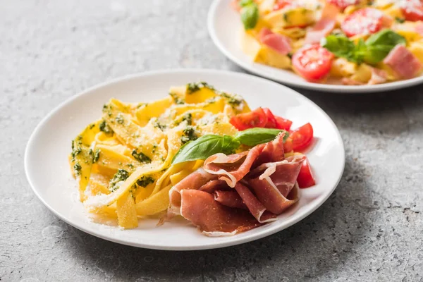 Selective focus of cooked Pappardelle with tomatoes, basil and prosciutto near ingredients on grey surface — Stock Photo
