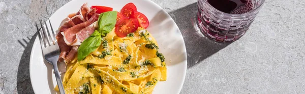 Top view of tasty Pappardelle with tomatoes, pesto and prosciutto with fork near red wine on grey surface, panoramic shot — Stock Photo