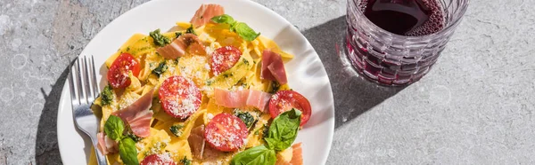 Vista superior de Pappardelle saboroso com tomates, pesto e prosciutto com garfo perto de vinho tinto na superfície cinza, tiro panorâmico — Fotografia de Stock