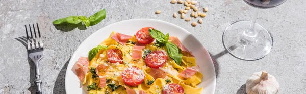 Pappardelle with tomatoes, pesto and prosciutto near red wine and ingredients on grey surface, panoramic shot — Stock Photo