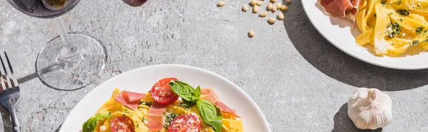 Fresh Pappardelle with tomatoes, pesto and prosciutto near red wine and ingredients on grey surface, panoramic shot — Stock Photo