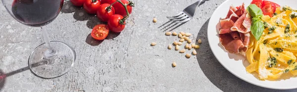 Pappardelle fraîche aux tomates, au pesto et au prosciutto près du vin rouge et des ingrédients à la surface grise, plan panoramique — Photo de stock