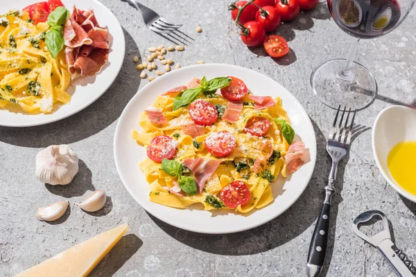 Pappardelle fresche con pomodori, pesto e prosciutto vicino al vino rosso e ingredienti sulla superficie grigia — Foto stock