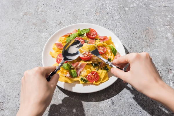 Cropped view of woman eating tasty Pappardelle with tomatoes, pesto and prosciutto with fork and spoon on grey surface — Stock Photo