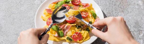 Cropped view of woman eating tasty Pappardelle with tomatoes, pesto and prosciutto with fork and spoon on grey surface, panoramic shot — Stock Photo