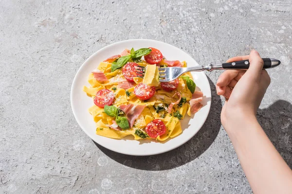 Cropped view of woman eating tasty Pappardelle with tomatoes, pesto and prosciutto with fork on grey surface — Stock Photo