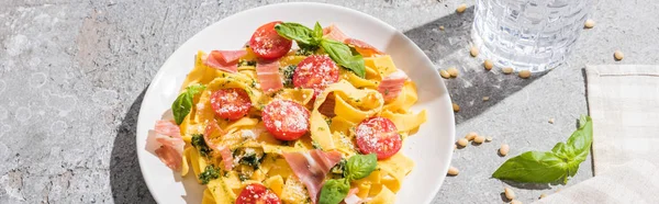 Tasty Pappardelle with tomatoes, pesto and prosciutto near water, napkin and pine nuts on grey surface, panoramic shot — Stock Photo