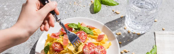 Partial view of woman eating tasty Pappardelle with tomatoes, pesto and prosciutto near water on grey surface, panoramic shot — Stock Photo