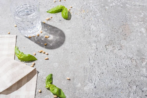 Basil leaves near water, napkin and pine nuts on grey surface — Stock Photo