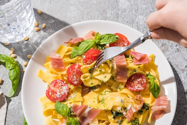 Partial view of woman eating tasty Pappardelle with tomatoes, pesto and prosciutto near water on grey surface — Stock Photo