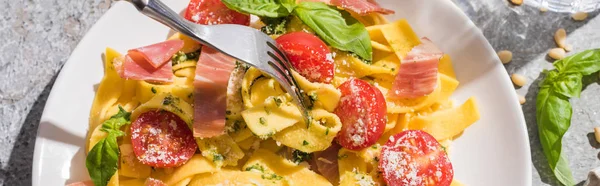 Top view of tasty Pappardelle with tomatoes, pesto and prosciutto with fork on grey surface, panoramic shot — Stock Photo