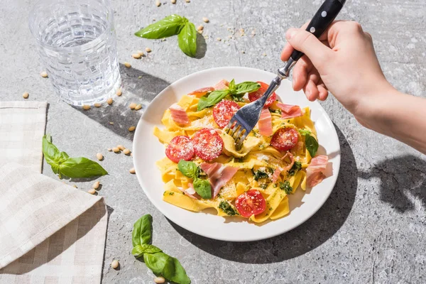 Visão parcial da mulher comendo pappardelle saboroso com tomates, pesto e prosciutto perto de água na superfície cinza — Fotografia de Stock