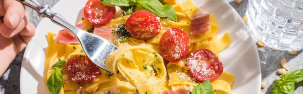 Visão parcial da mulher comendo pappardelle saboroso com tomates, pesto e prosciutto perto de água na superfície cinza, tiro panorâmico — Fotografia de Stock