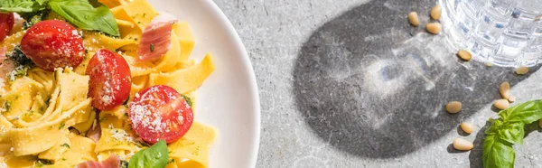 Top view of tasty Pappardelle with tomatoes, pesto and prosciutto near water on grey surface, panoramic shot — Stock Photo