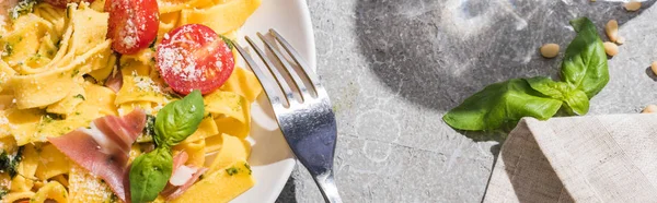 Top view of tasty Pappardelle with tomatoes, pesto and prosciutto with fork on grey surface, panoramic shot — Stock Photo