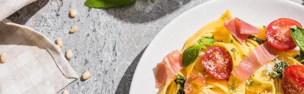 Top view of tasty Pappardelle with tomatoes, pesto and prosciutto near napkin and pine nuts on grey surface, panoramic shot — Stock Photo