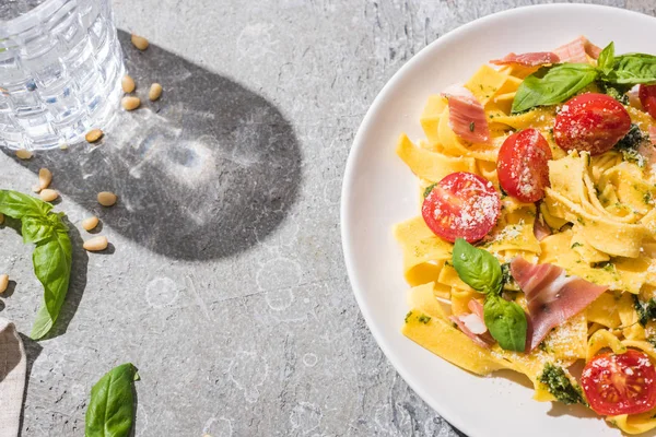 Top view of tasty Pappardelle with tomatoes, pesto and prosciutto near water on grey surface — Stock Photo