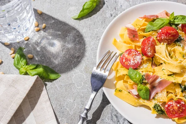 Top view of tasty Pappardelle with tomatoes, pesto and prosciutto with fork near water and pine nuts on grey surface — Stock Photo