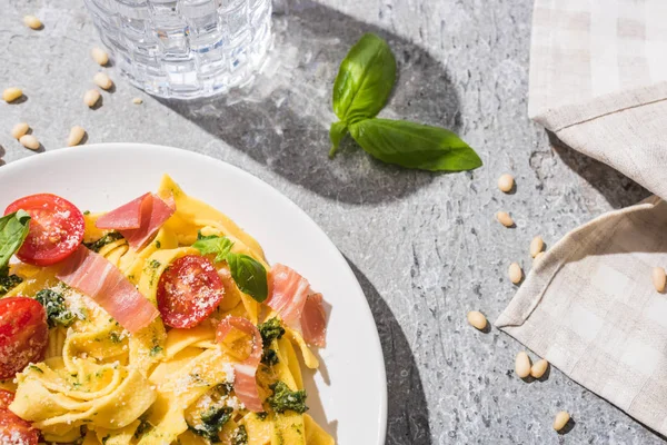 Top view of tasty Pappardelle with tomatoes, pesto and prosciutto near water and pine nuts on grey surface — Stock Photo