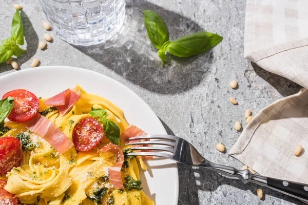 Blick von oben auf schmackhafte Pappardelle mit Tomaten, Pesto und Prosciutto mit Gabel am Wasser und Pinienkernen auf grauer Oberfläche — Stockfoto