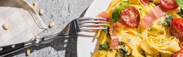 Top view of tasty Pappardelle with tomatoes, pesto and prosciutto with fork near pine nuts and napkin on grey surface, panoramic shot — Stock Photo