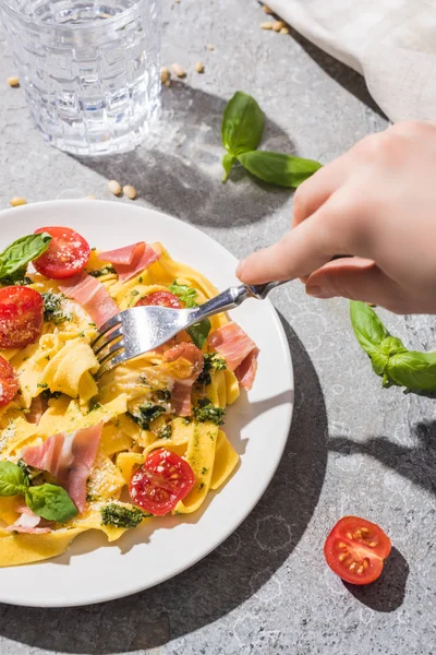 Visão parcial da mulher comendo pappardelle saboroso com tomates, pesto e prosciutto perto de água na superfície cinza — Fotografia de Stock