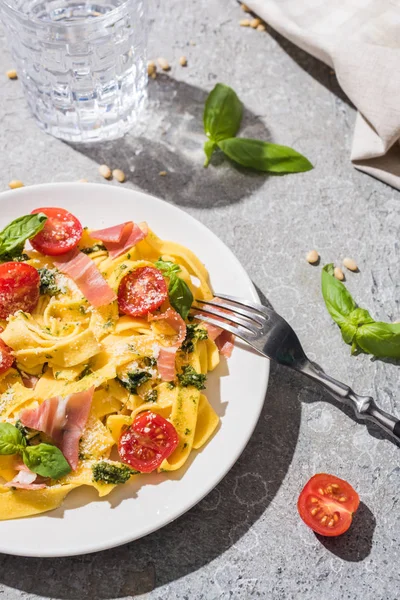 Tasty Pappardelle with tomatoes, pesto and prosciutto with fork near water and pine nuts on grey surface — Stock Photo