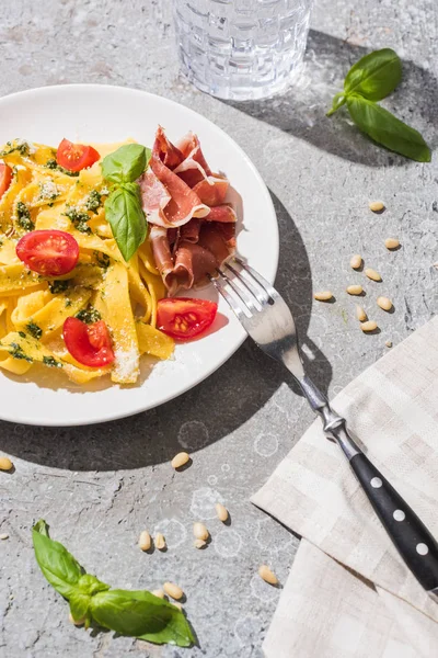 Tasty Pappardelle with tomatoes, pesto and prosciutto with fork near water and pine nuts on grey surface — Stock Photo