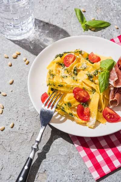 Tasty Pappardelle with tomatoes, pesto and prosciutto with fork near water and pine nuts on grey surface — Stock Photo