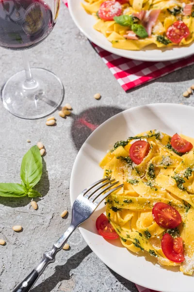 Tasty Pappardelle with tomatoes, pesto and prosciutto with fork near red wine, basil and pine nuts on grey surface — Stock Photo