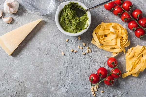 Top view of raw Pappardelle near tomatoes, garlic, pine nuts, water and pesto sauce on grey surface — Stock Photo