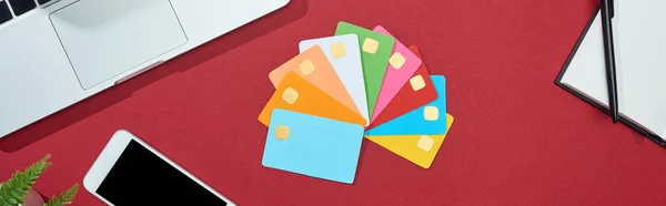 Top view of multicolored empty credit cards on red background with smartphone, laptop, earphones and coffee, panoramic shot — Stock Photo