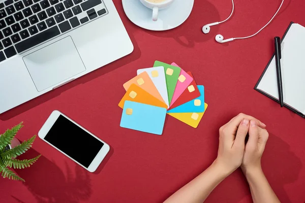 Частичный вид женщины рядом с многоцветными пустыми кредитными картами на красном фоне со смартфоном, ноутбуком, наушниками и кофе — стоковое фото