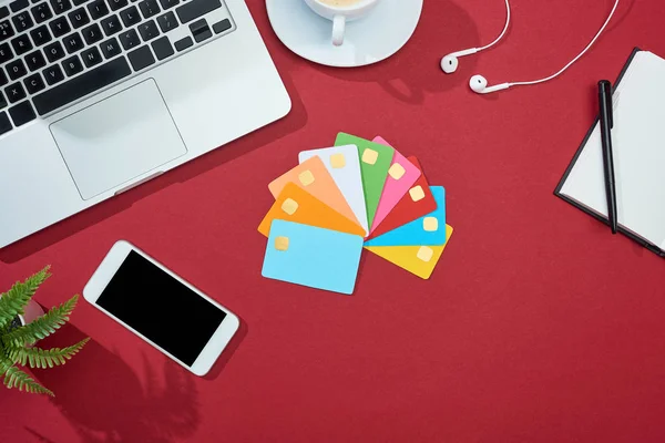 Ansicht von bunten leeren Kreditkarten auf rotem Hintergrund mit Smartphone, Laptop, Kopfhörer und Kaffee — Stockfoto