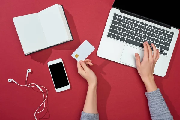 Vista superior do cartão de crédito no fundo vermelho com smartphone, laptop, fones de ouvido, café, notebook e planta — Fotografia de Stock