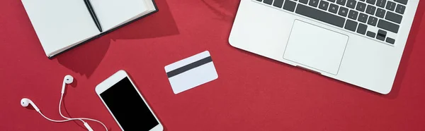 Top view of credit card on red background with smartphone, laptop, earphones, notebook, panoramic shot — Stock Photo