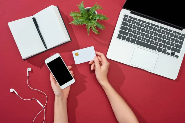 Cropped view of woman holding credit card and smartphone near laptop, earphones, pen, notebook and plant on red background — Stock Photo