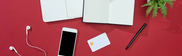 Vista superior do cartão de crédito, smartphone, laptop, fones de ouvido, caneta, notebook e planta no fundo vermelho, tiro panorâmico — Fotografia de Stock