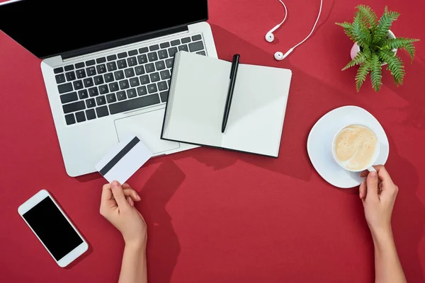 Partial view of woman holding credit card and cup of coffee near smartphone, laptop, earphones, notebook and plant on red background — Stock Photo